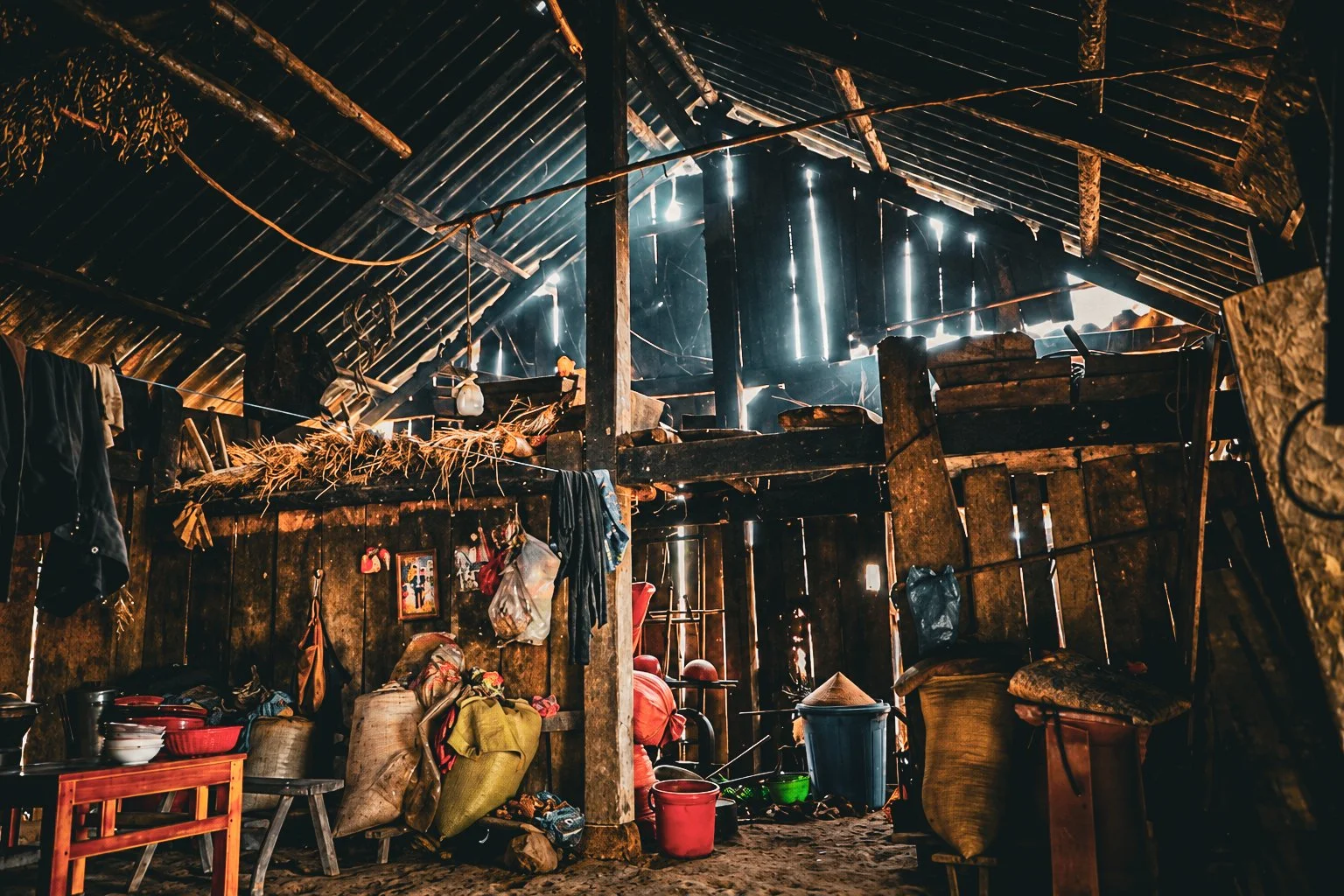 Hmong house interior showing timber posts, a storage loft, baskets and household utensils beneath a high roof.
