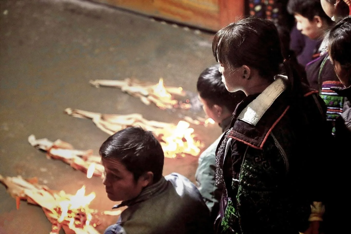 Community gathers indoors as rows of paper offerings burn on the floor during a Hmong household ritual.