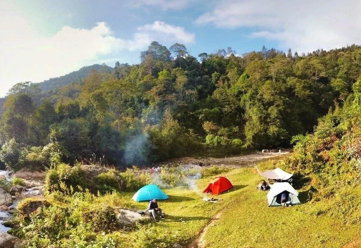Camping scene with three tents on grassy area near a small stream, surrounded by trees and hills under a partly cloudy sky.