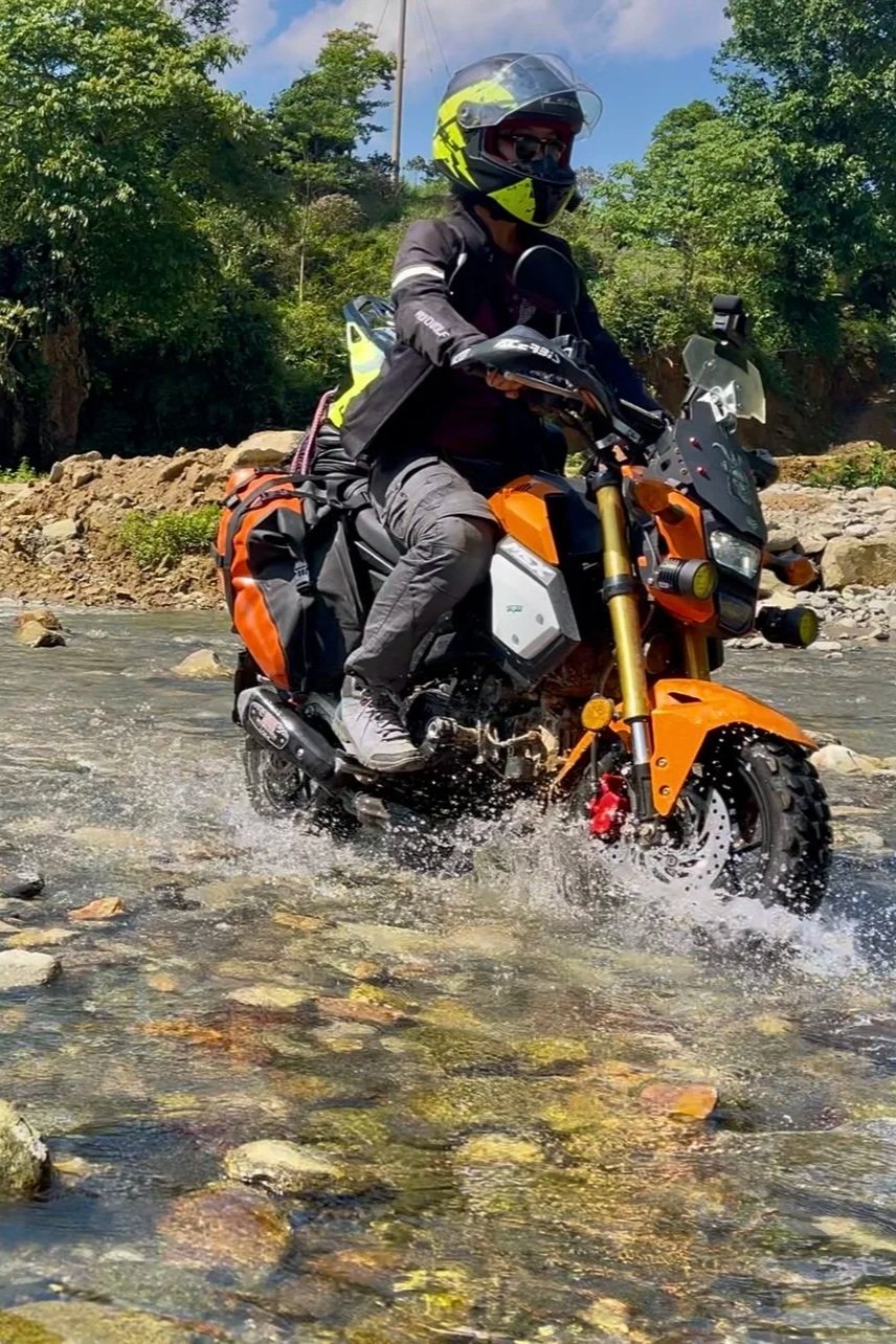 Motorcycle rider crossing a shallow river with trees and clear sky in the background.