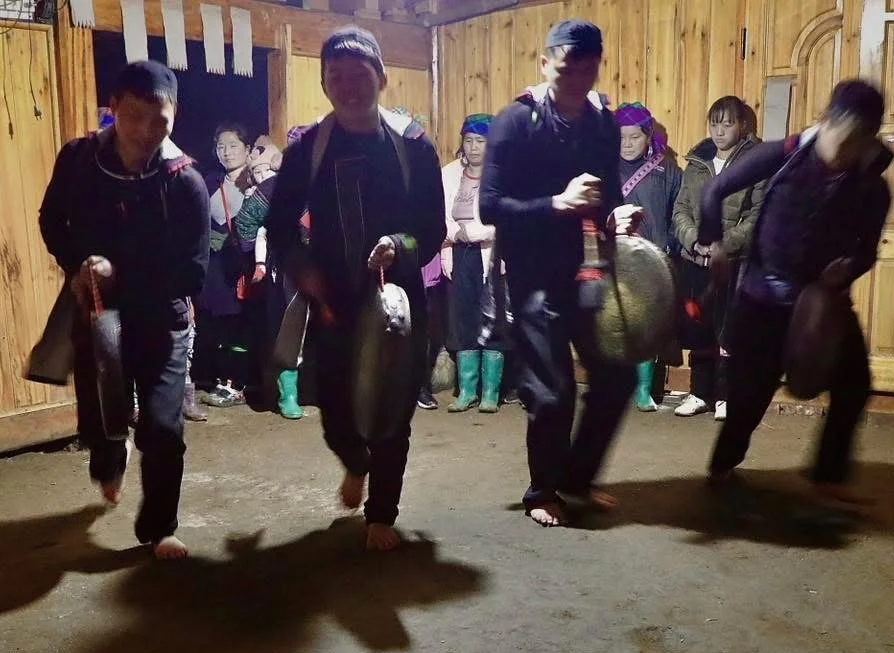 Group of Hmong men step in unison while carrying gongs as villagers watch an indoor celebration.