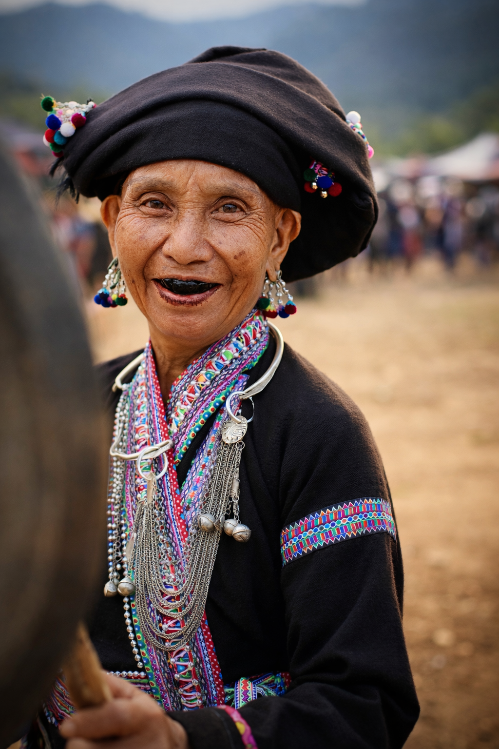 A Lao woman in traditional dress adorned with layered silver jewellery and colourful embroidery, smiling during Bun Vốc Nặm as community life unfolds behind her.