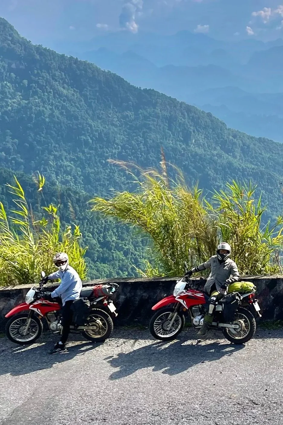 Two motorcyclists wearing helmets and jackets, parked on a mountain road with a lush green landscape and misty mountains in the background.