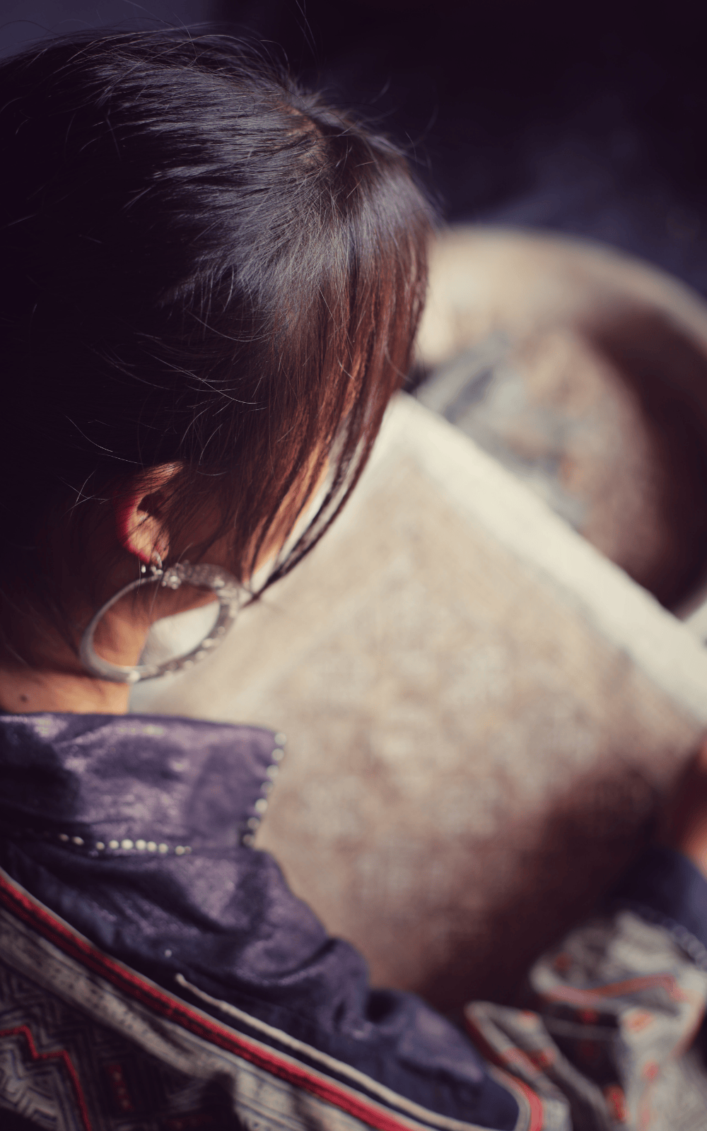 Over-shoulder view of a Hmong woman focused on textile work, wearing tradtional clothing while crafting patterned fabric by hand.