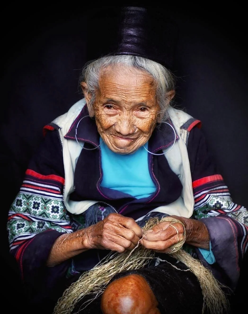 Elderly Hmong woman named May sitting and weaving natural fibers with her hands while wearing traditional clothing.