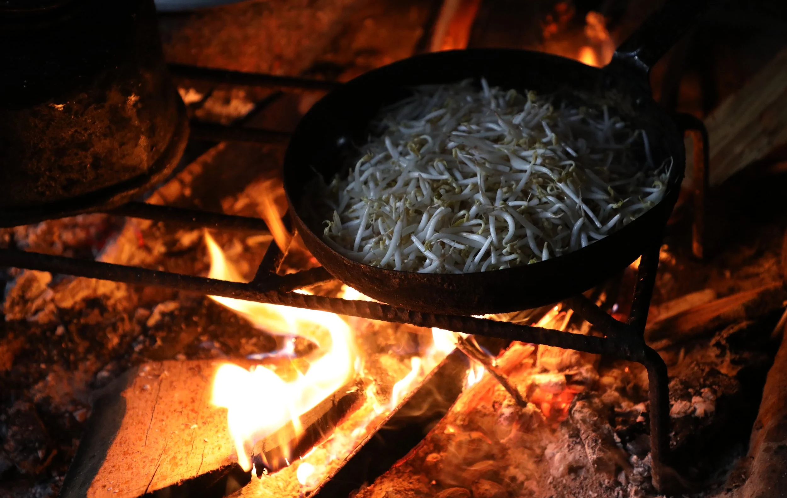 Food cooking in a shallow black pan supported above burning logs in a Hmong household hearth.