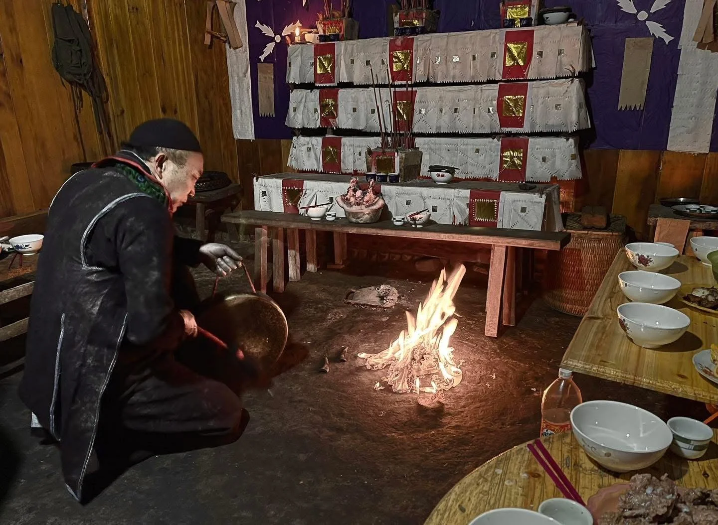 Hmong shaman kneels by a small hearth, praying before an altar covered with paper charms, bowls, and offerings.