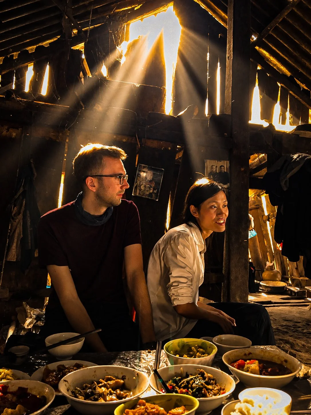 Two people seated beside a shared meal inside a Hmong timber home, with shafts of sunlight entering through the upper walls.