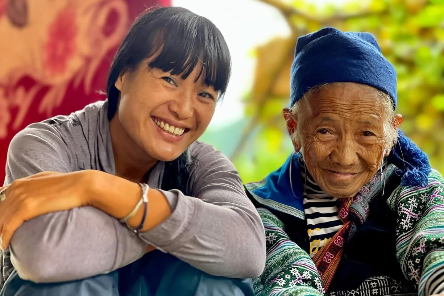 Elderly Hmong woman May smiling beside a younger woman, both sitting close together and sharing a warm moment.