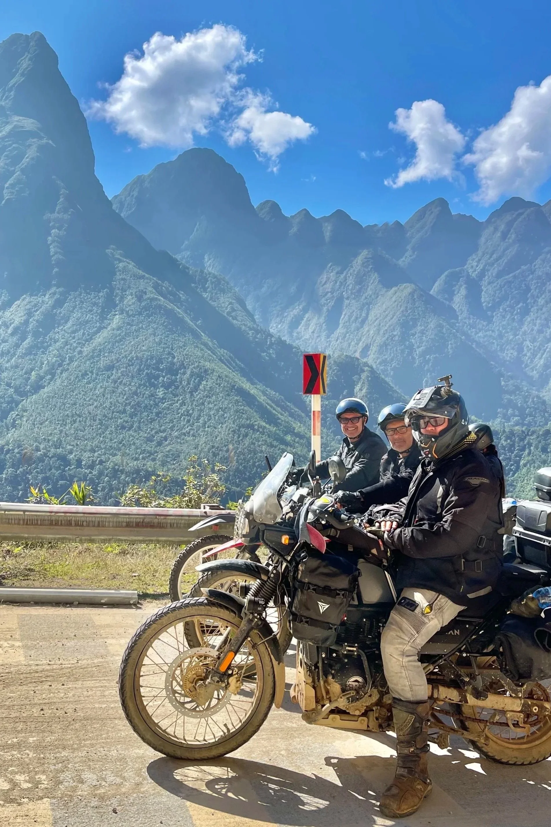 Three motorcycle riders in gear and helmets pose on their bikes, with a backdrop of green mountains and a blue sky with clouds.