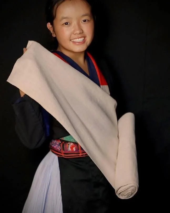 Hmong woman wearing traditional clothing and holding a large roll of natural hemp fabric used for weaving