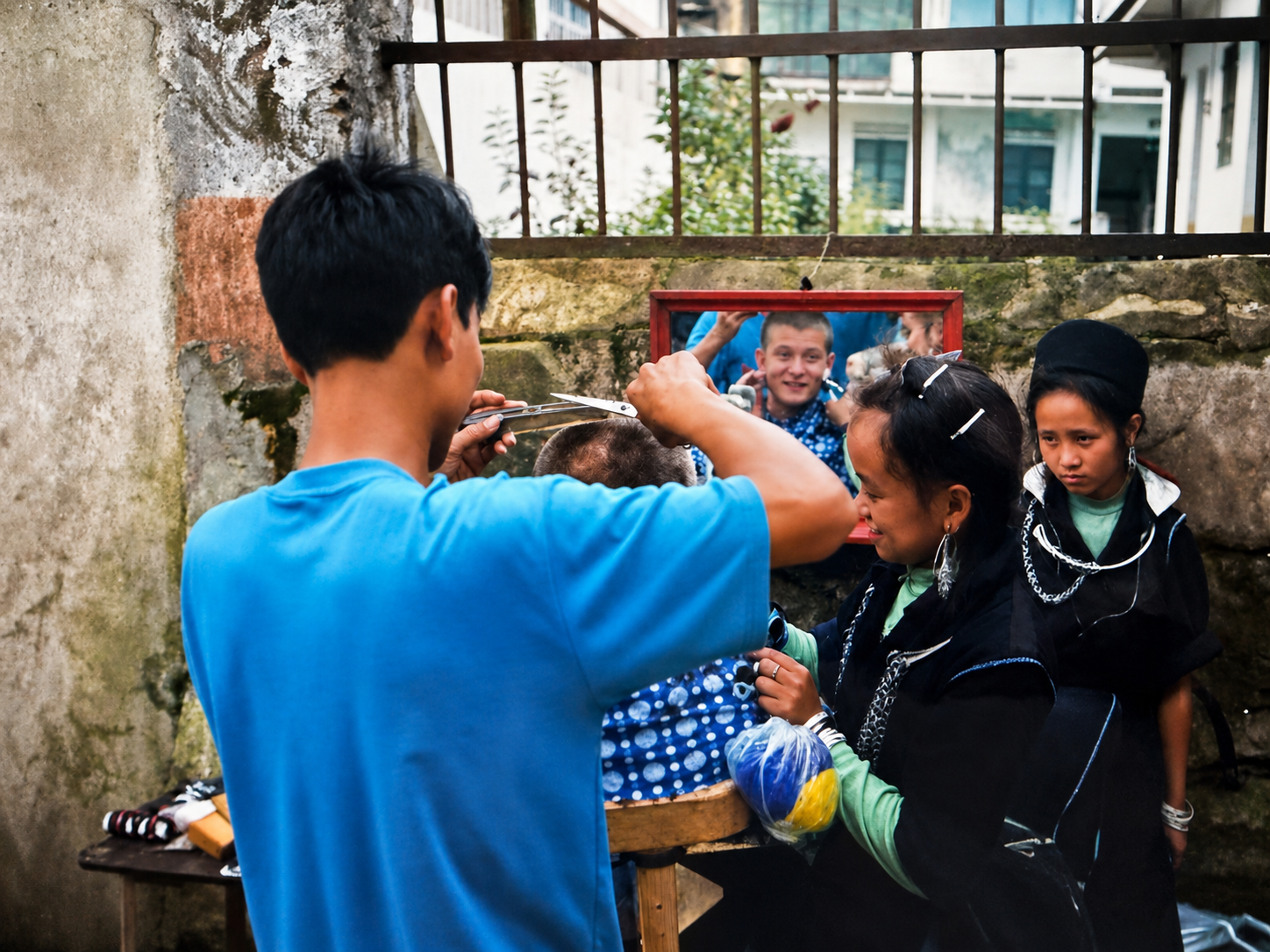 Street barber cutting a colleague's hair on the market steps in Sapa, Vietnam, in 1998. Hmong girls spectate and later participate in the trim.