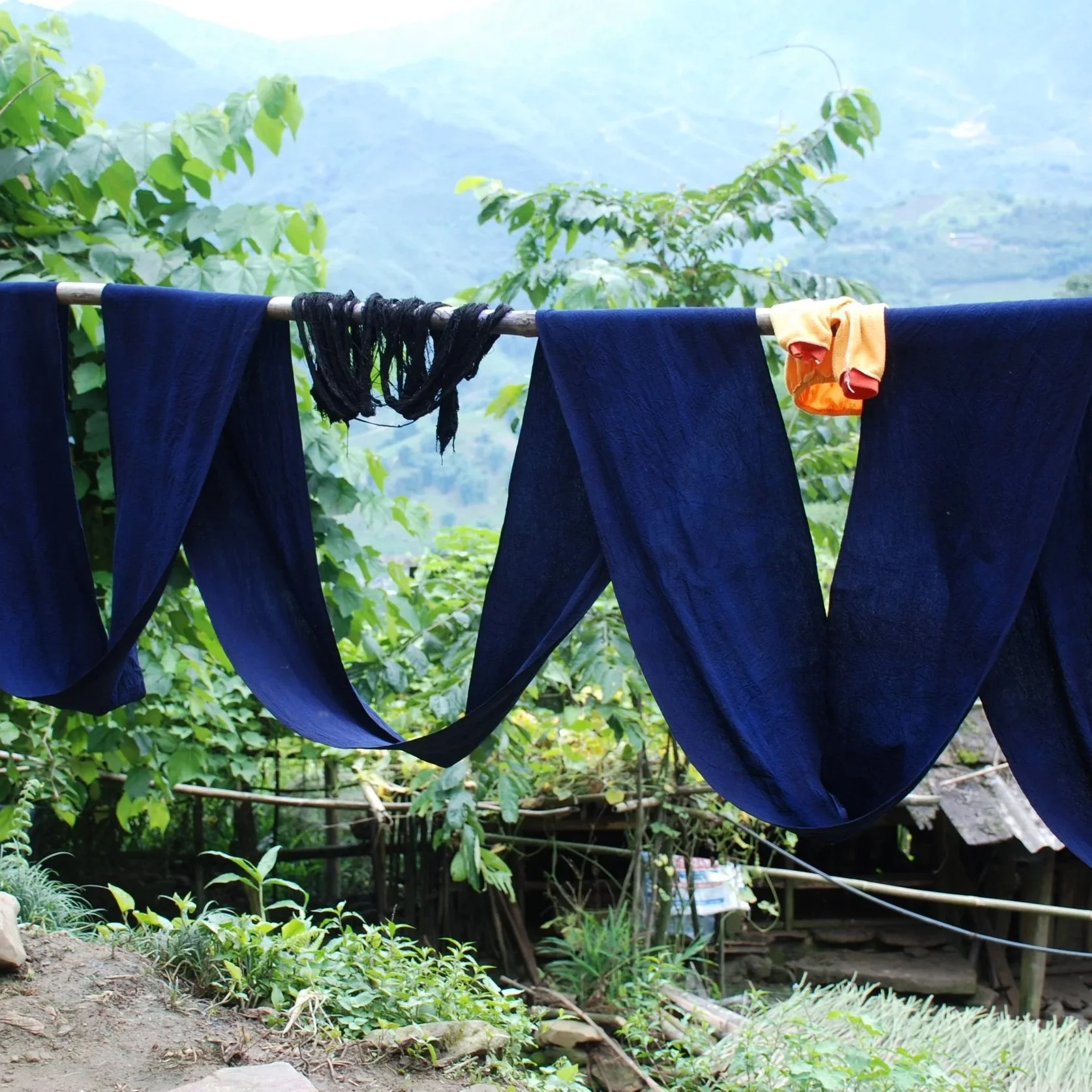 Long lengths of dark indigo cloth draped over a pole to dry above a village house, with mountains beyond.