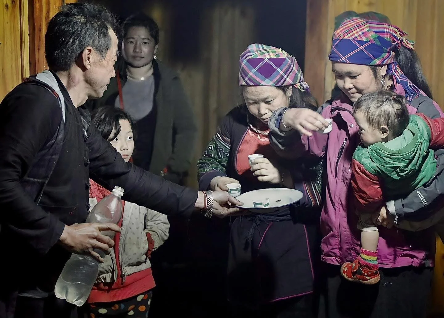 Family receives small cups of rice wine during a Hmong blessing inside the home; women and children stand nearby.