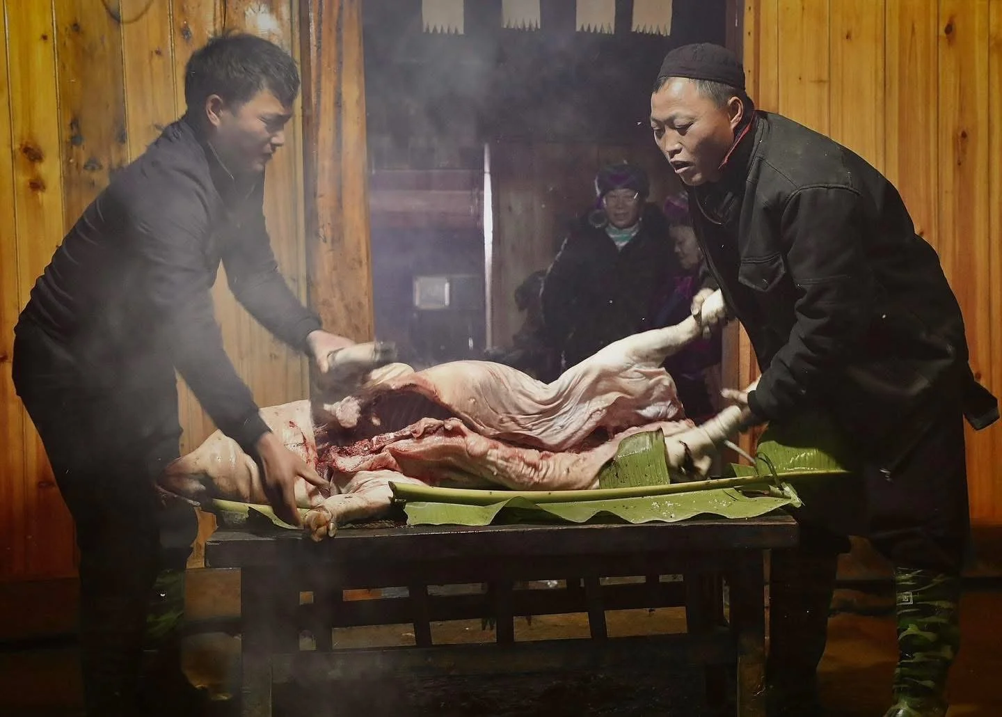 Two men prepare a freshly slaughtered pig on a table for a Hmong New Year feast inside a wooden house.
