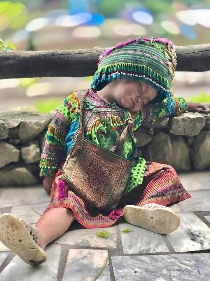 A small Hmong child wearing traditional green and embroidered clothing sleeps while seated on a stone path in a mountain village, leaning gently against a low wall.