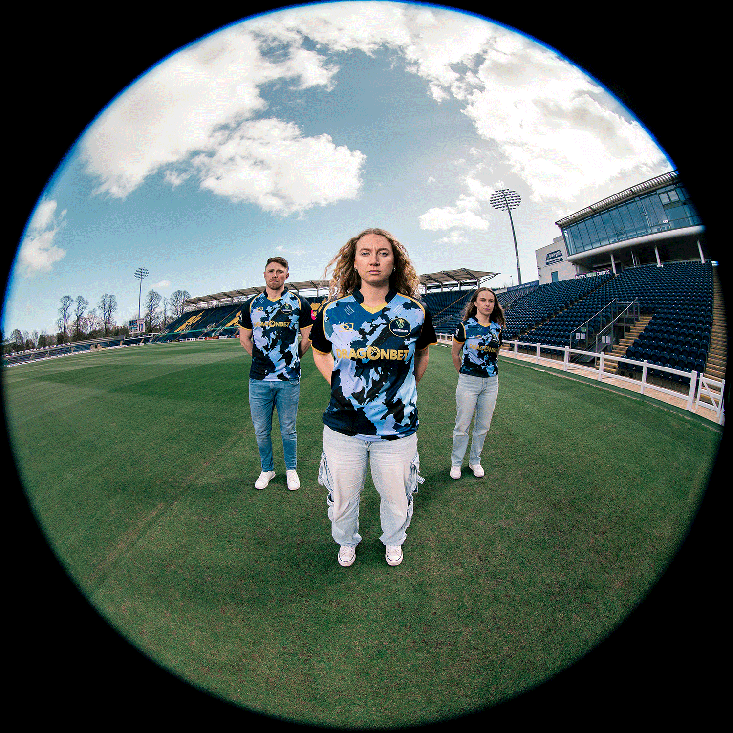Three young women standing on a cricket field, wearing blue camouflage jerseys with orange logos, with empty stadium seating and a cloudy sky in the background.