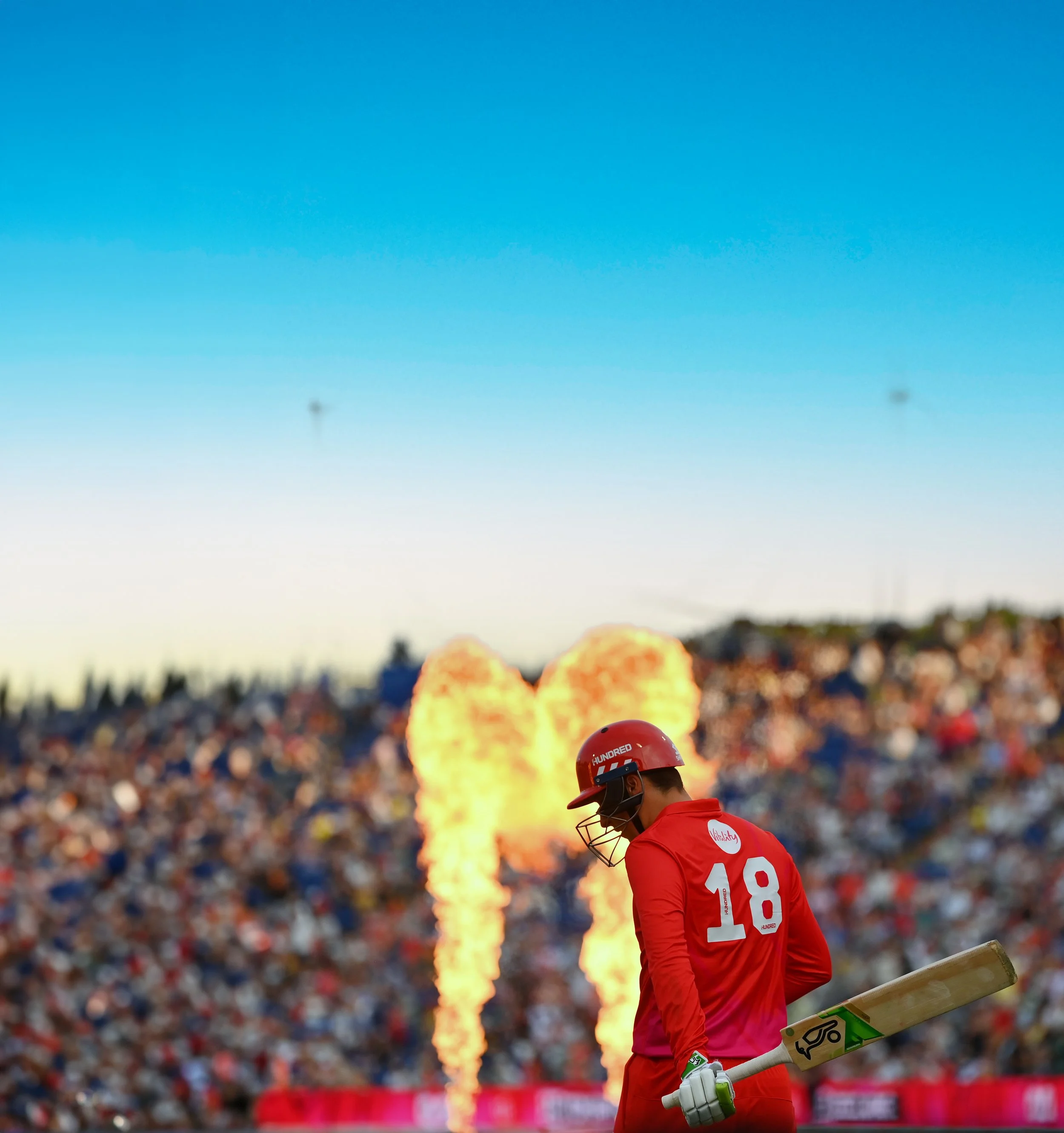 A cricket player in red uniform holding a bat, with flames and a large crowd in the background during a cricket match.