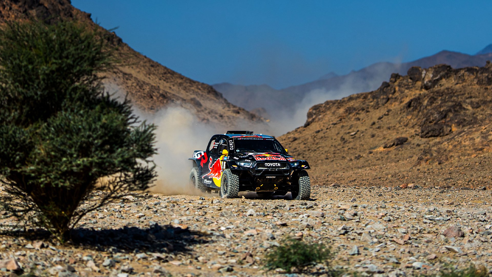 Dakar Rally rally car racing on a dusty, rocky desert trail with mountains in the background.