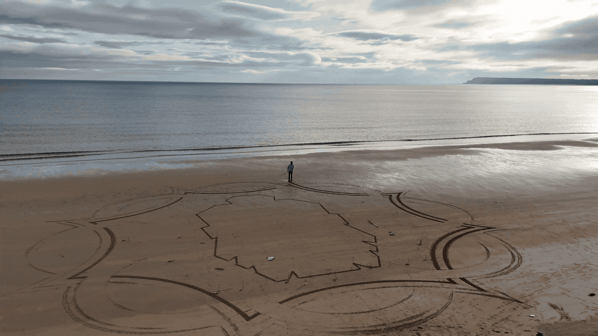 A person at the beach stands in front of a large geometric sand art design that resembles the outline of a country, with roads or paths radiating from it, and the ocean and cloudy sky in the background.