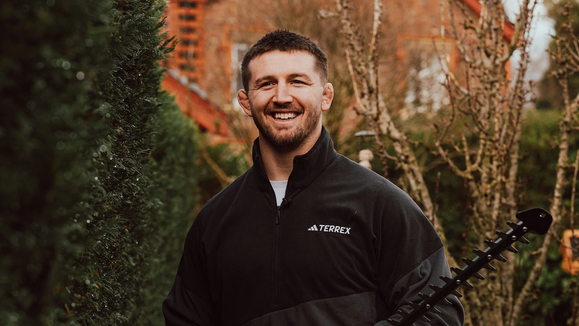 Tom Curry, English Rugby, wearing a black Terrex jacket, standing outdoors on a cloudy day surrounded by trees and bushes, with a red-brick building in the background. Stiga Gardening.