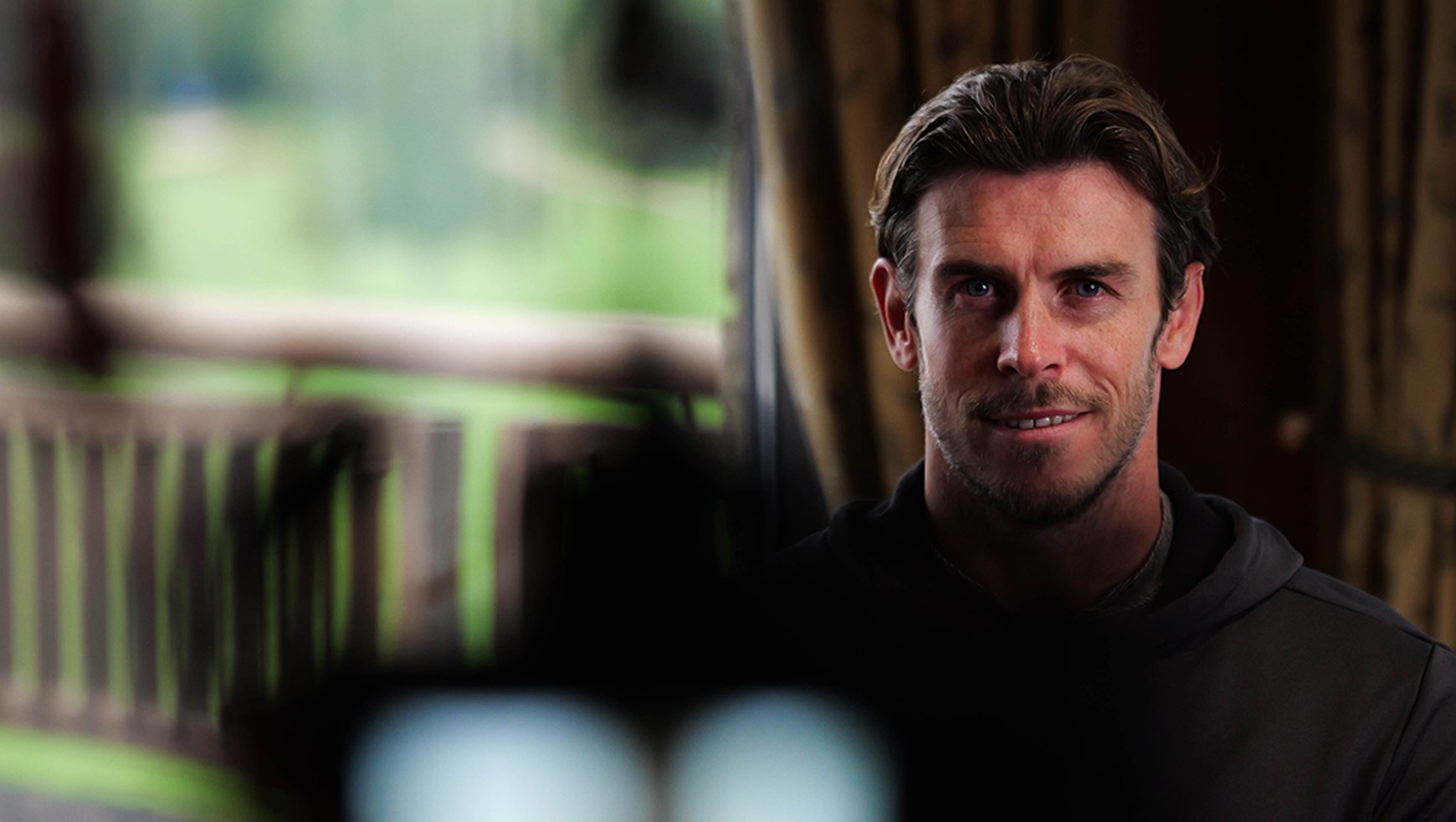 A man with dark hair and light skin, smiling and looking at the camera, standing in a dimly lit indoor setting with blurred background and a window showing green foliage outside.