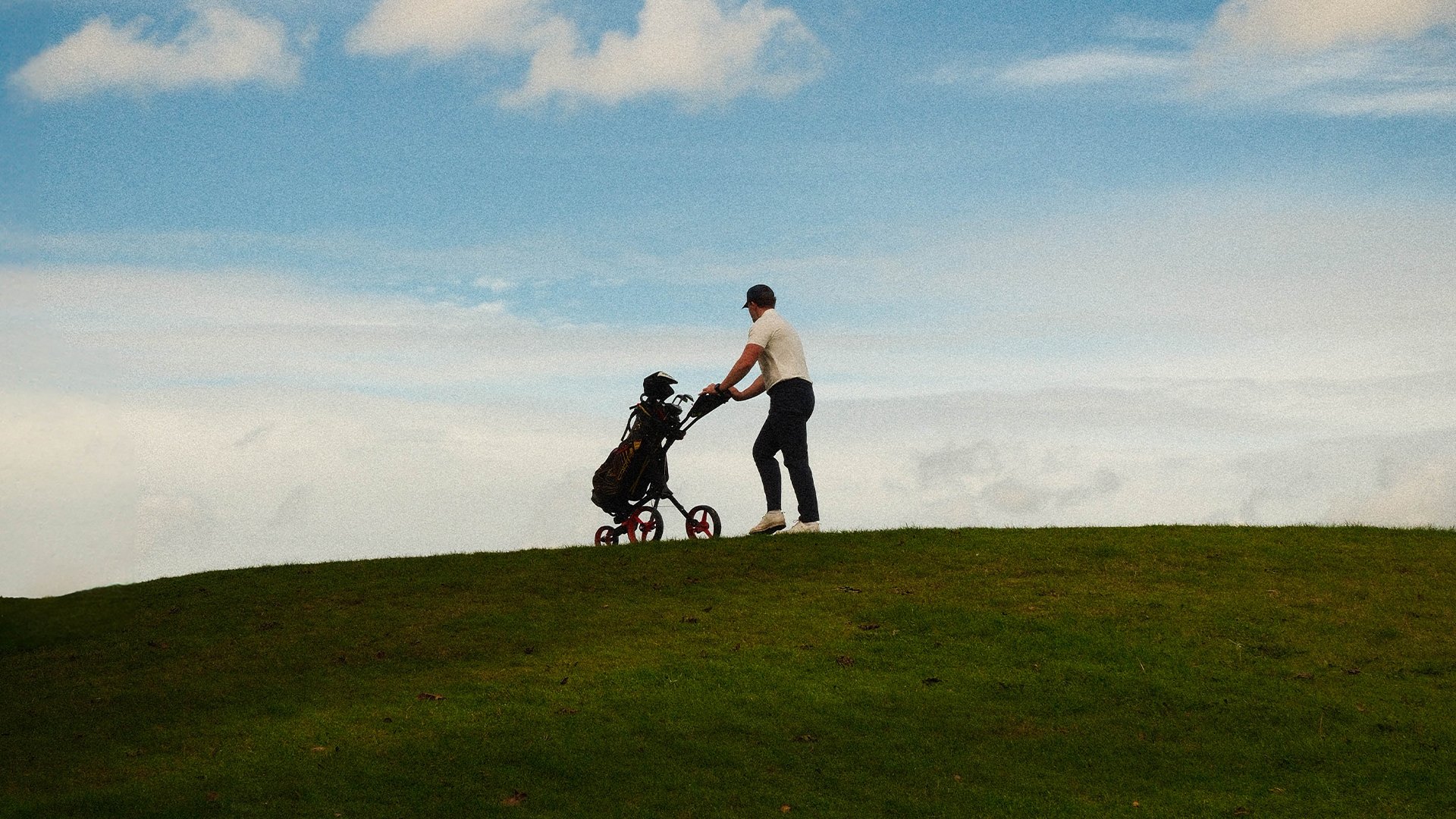 Bont Golf. A person in a white shirt, black pants, and white shoes standing next to a small red and black golf cart filled with golf clubs on a grassy hill under a partly cloudy sky.