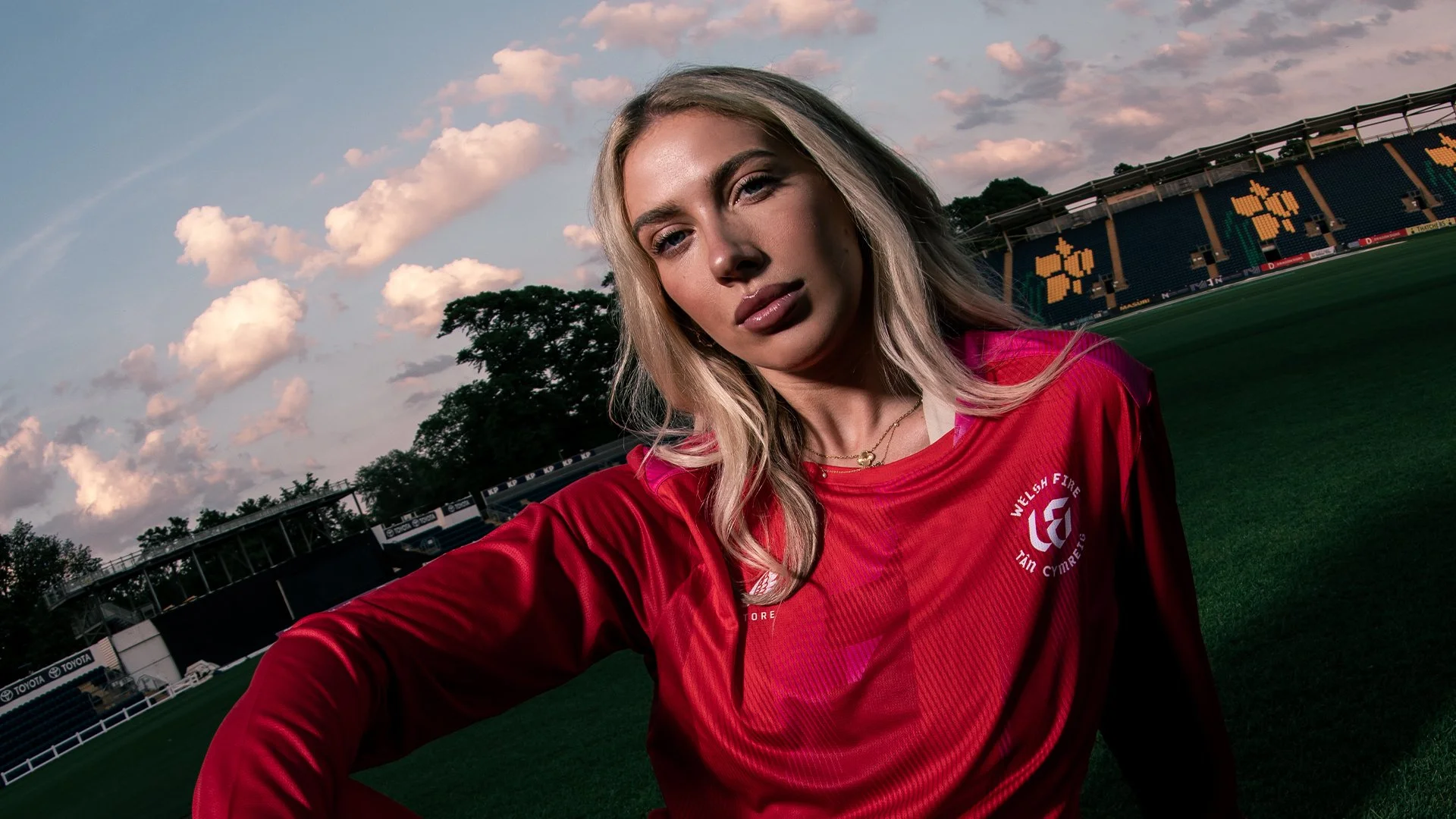 Sports photgraphy during sunset, wearing a red sports jersey with Welsh Fire logo, with stadium seating and trees in the background.