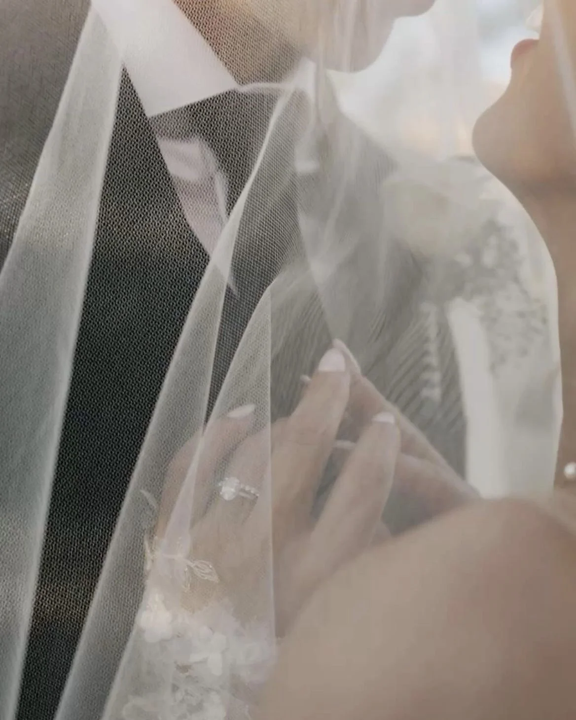 A bride and groom with a veil between them, focusing on their hands and faces, with the bride touching the groom's chest.