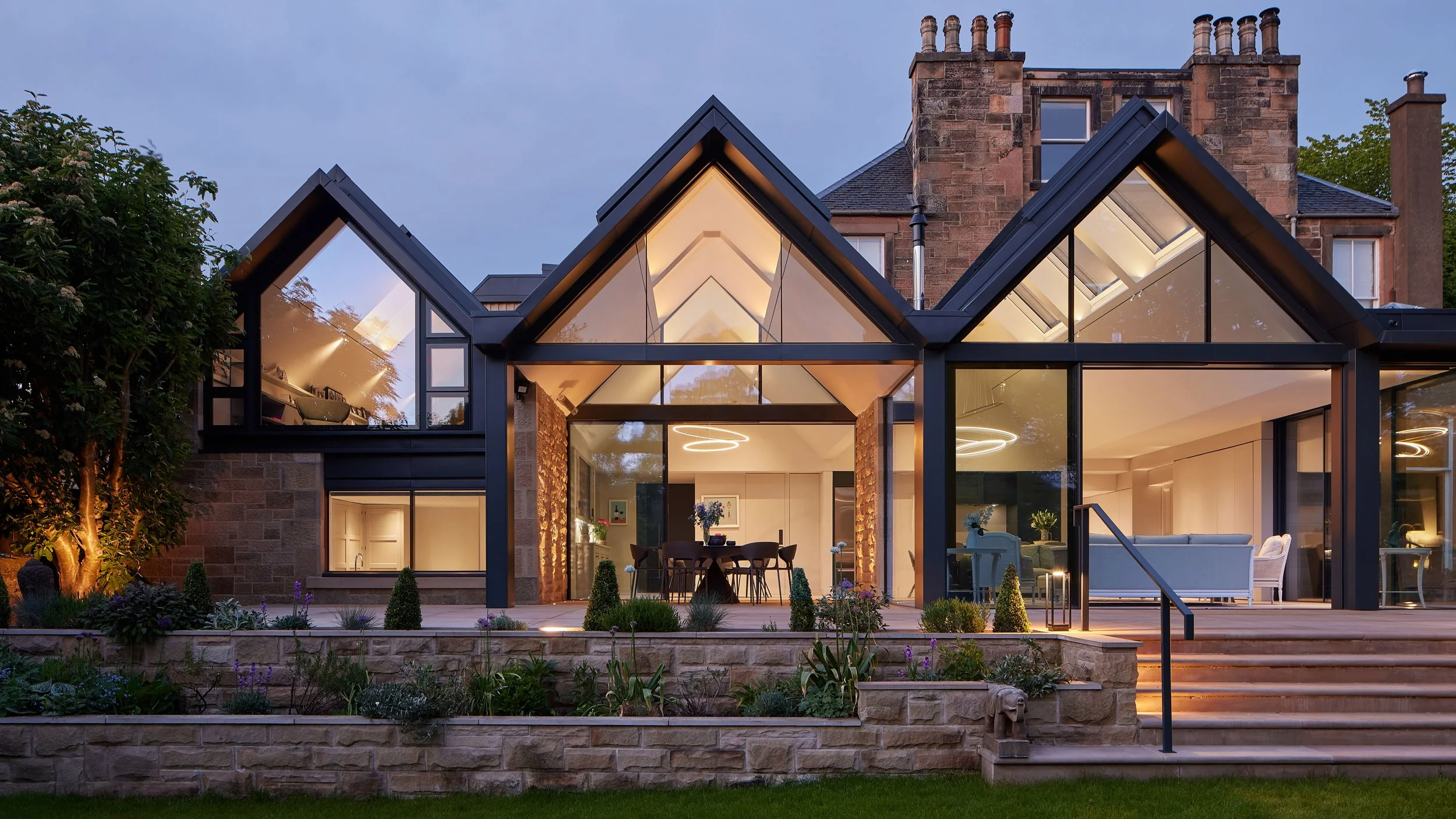 A modern house with glass walls and roof, illuminated from inside, with a stone and brick exterior, set in a garden during dusk.
