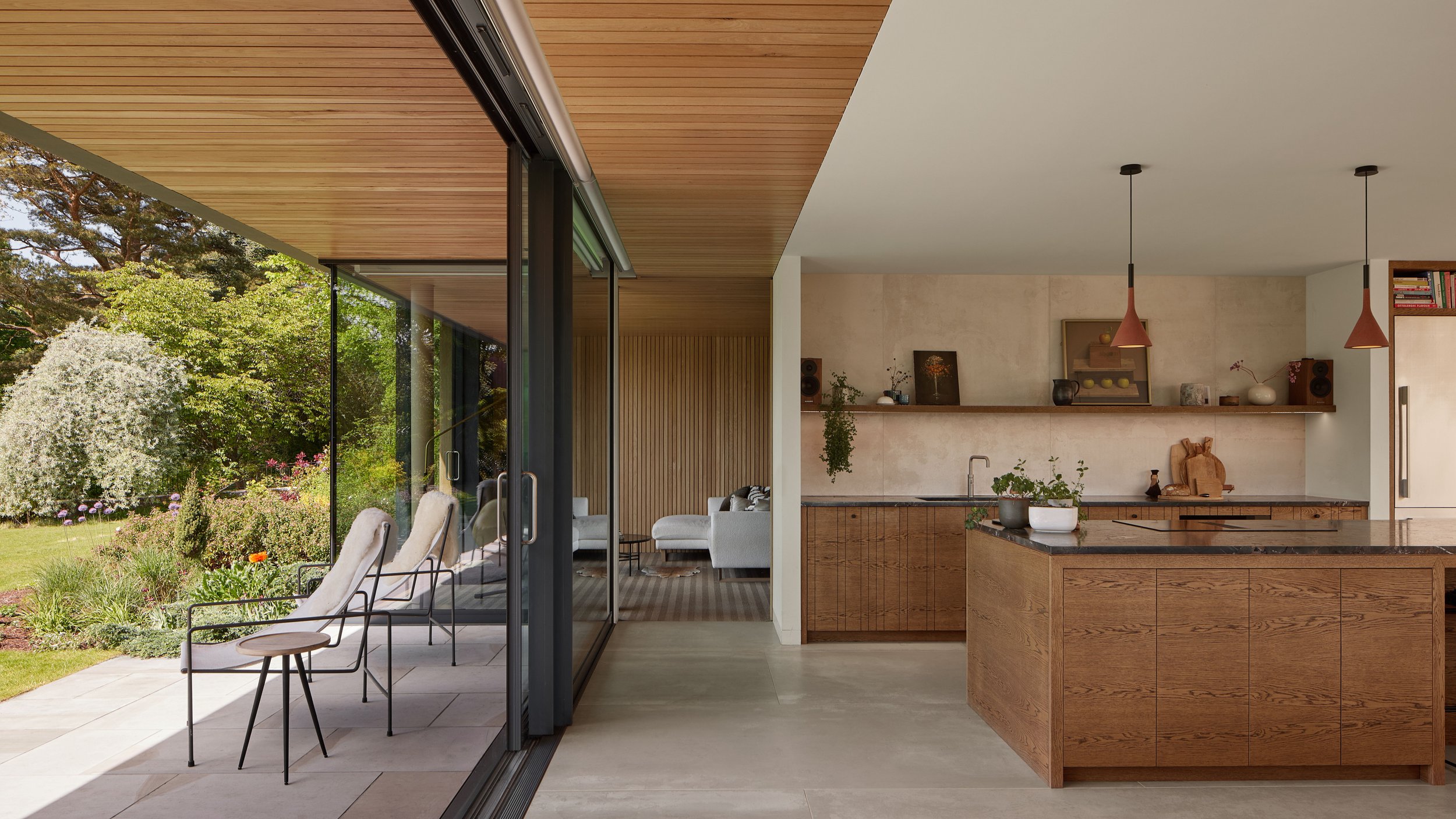 Modern kitchen with wooden cabinets and a black countertop, open to a patio with outdoor chairs and a garden view.