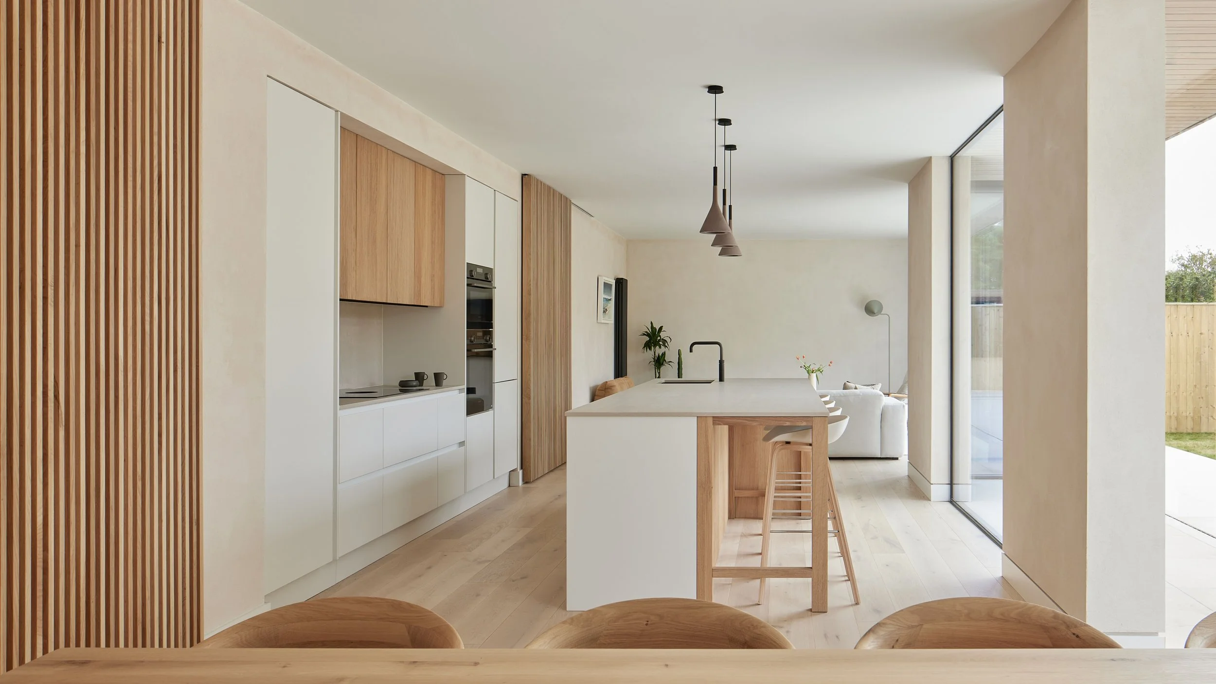 Modern open-concept kitchen and living room with minimalist design, white and wooden cabinetry, island with bar stools, and large sliding glass doors leading to an outdoor patio.