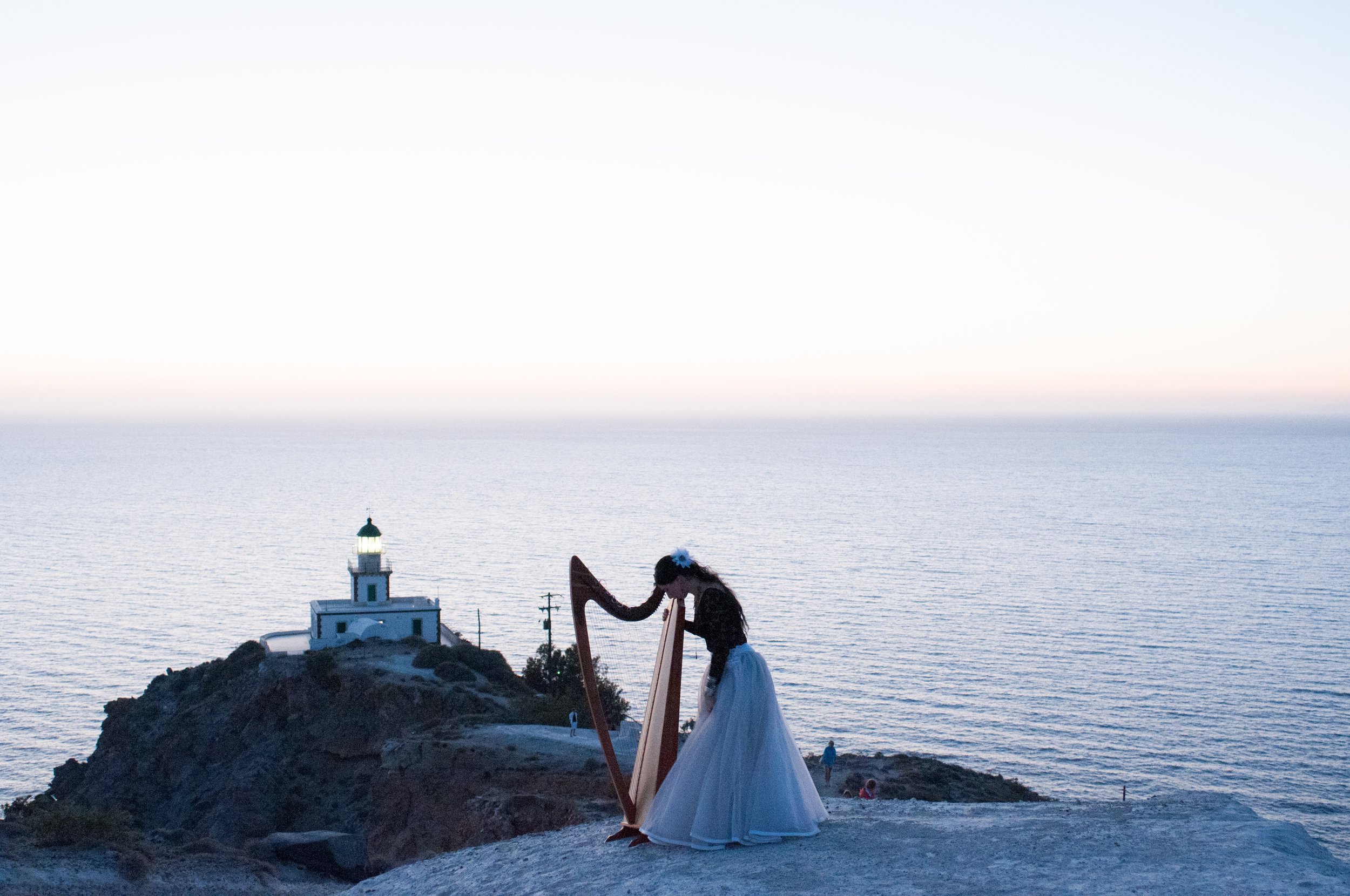 A coastal scene at sunset with a small lighthouse on a rocky outcrop, a woman in a white dress playing a harp, and a few people walking near the water.