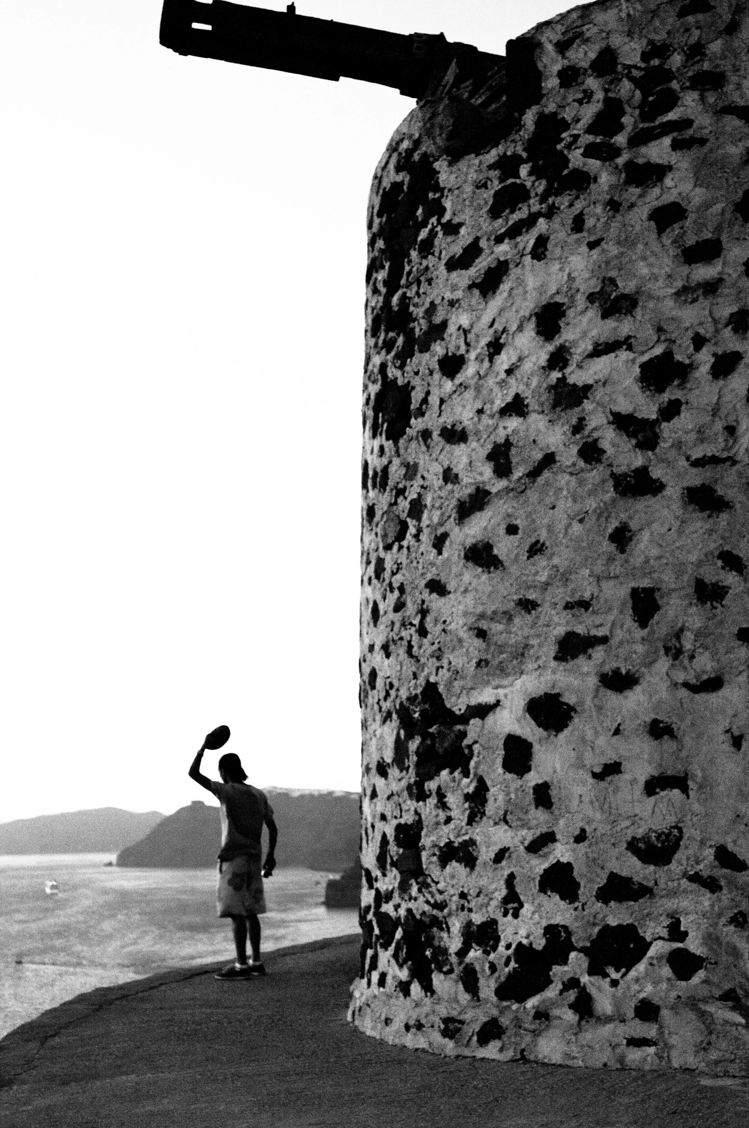 A person standing on a pathway by the ocean, with another person playing with a ball nearby, near a large, textured rock formation, during sunset or sunrise.