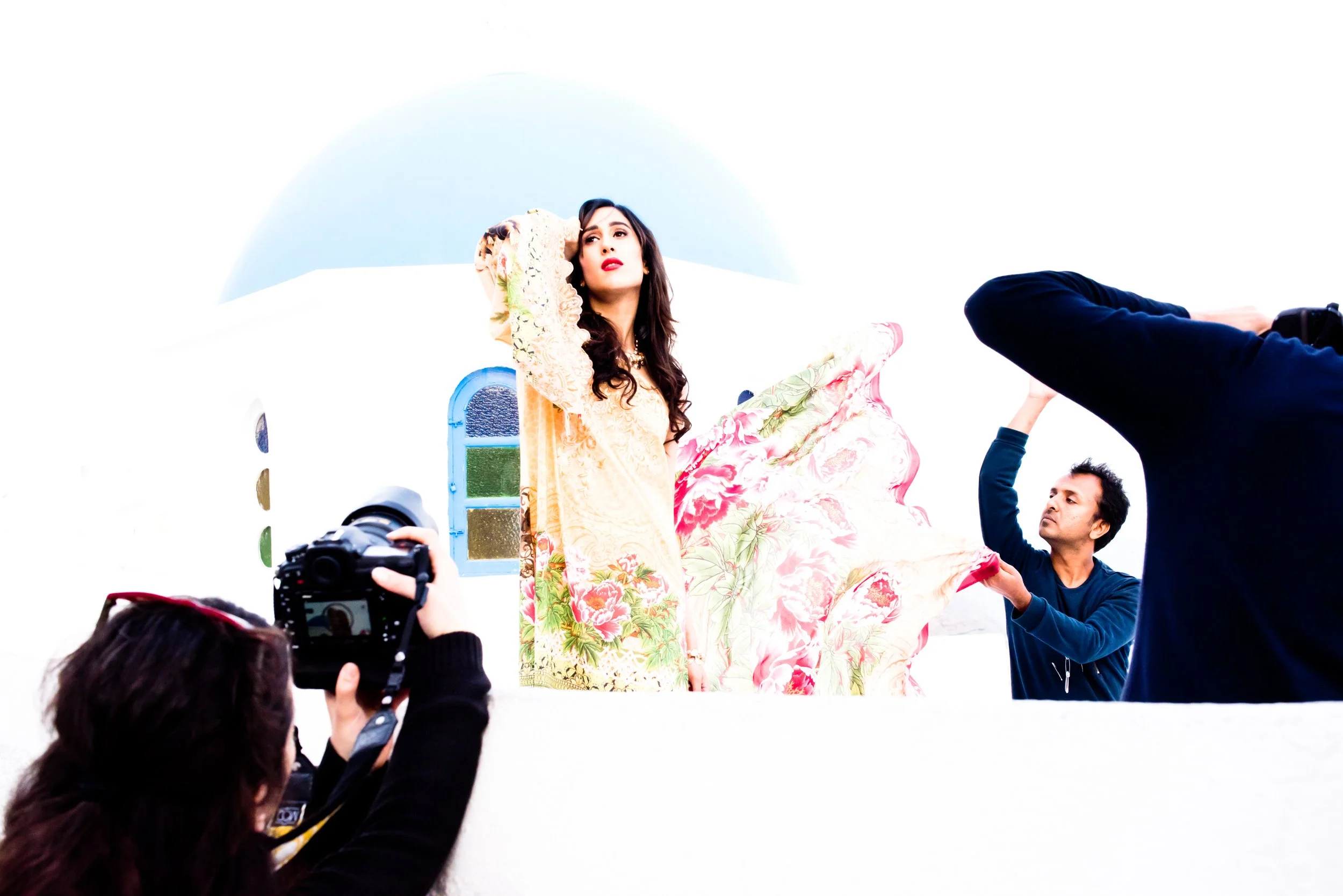 A woman in a floral saree is being photographed or filmed by crew members in an outdoor setting with a white background and a small arched blue window visible behind her.