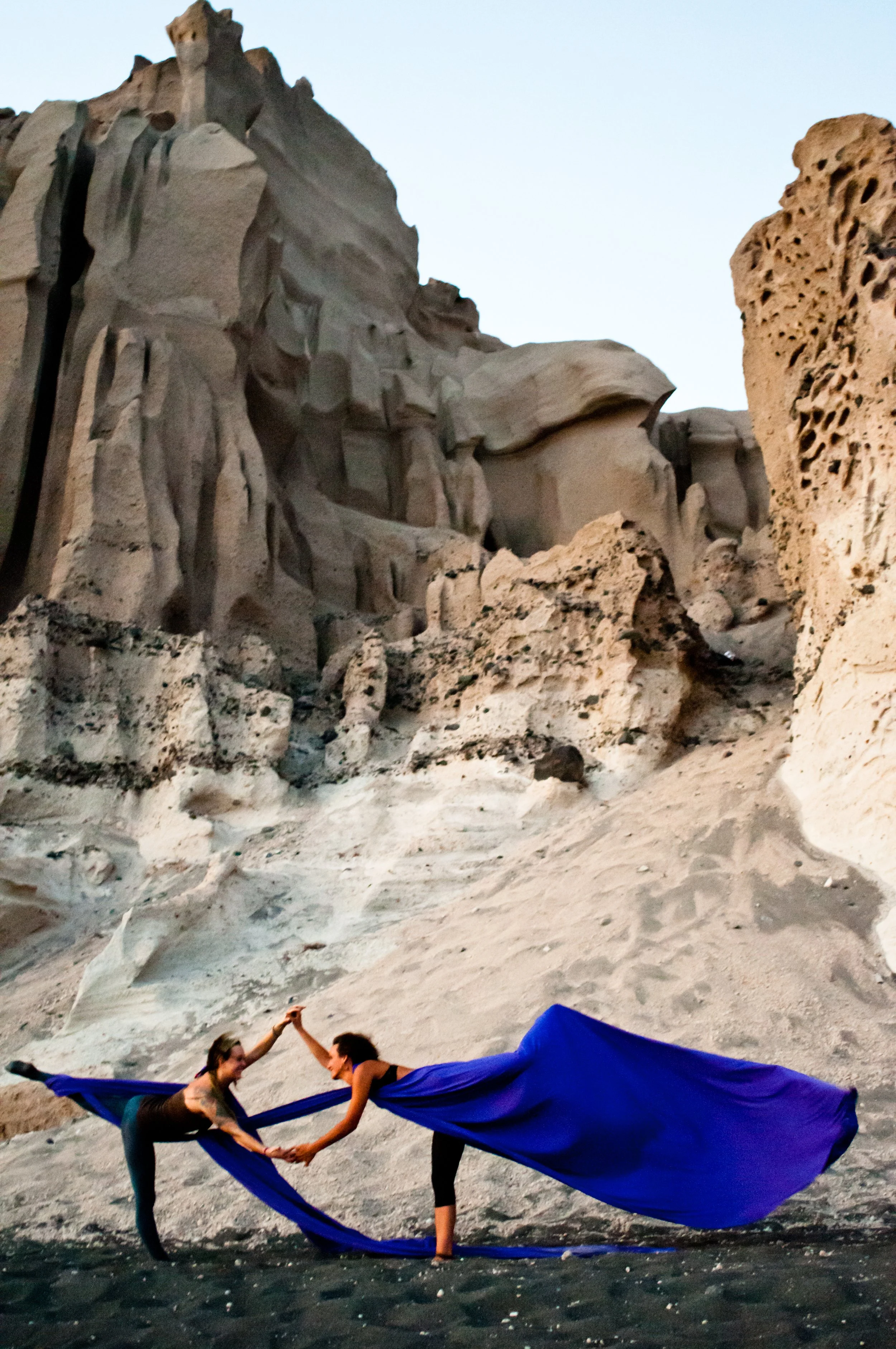 Two women are practicing acro yoga on a sandy beach, holding a blue fabric between them as part of their pose, with large rock formations in the background.