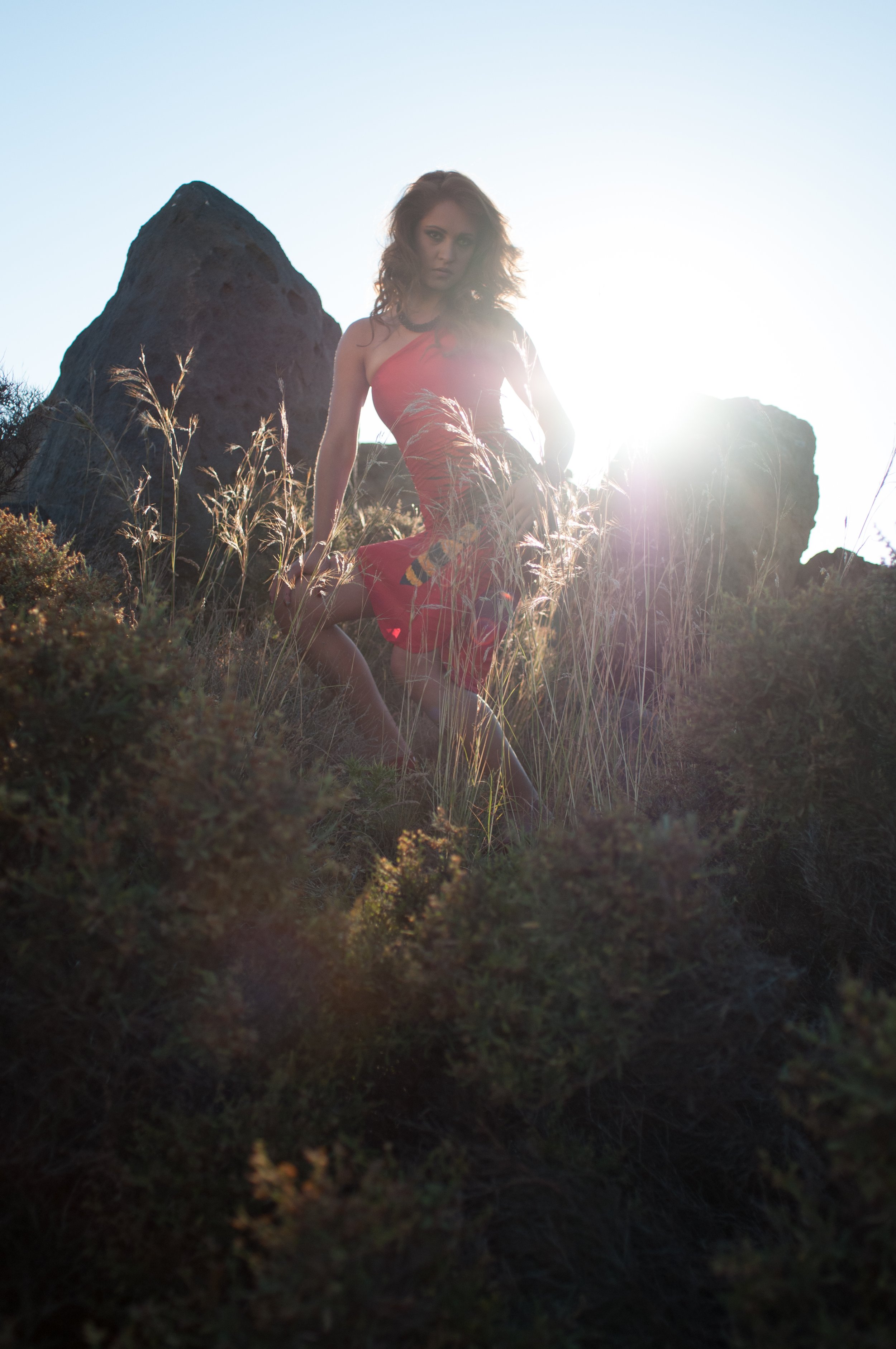 A woman with curly hair wearing a red dress kneeling in tall grass with large rocks behind her, backlit by the setting sun.