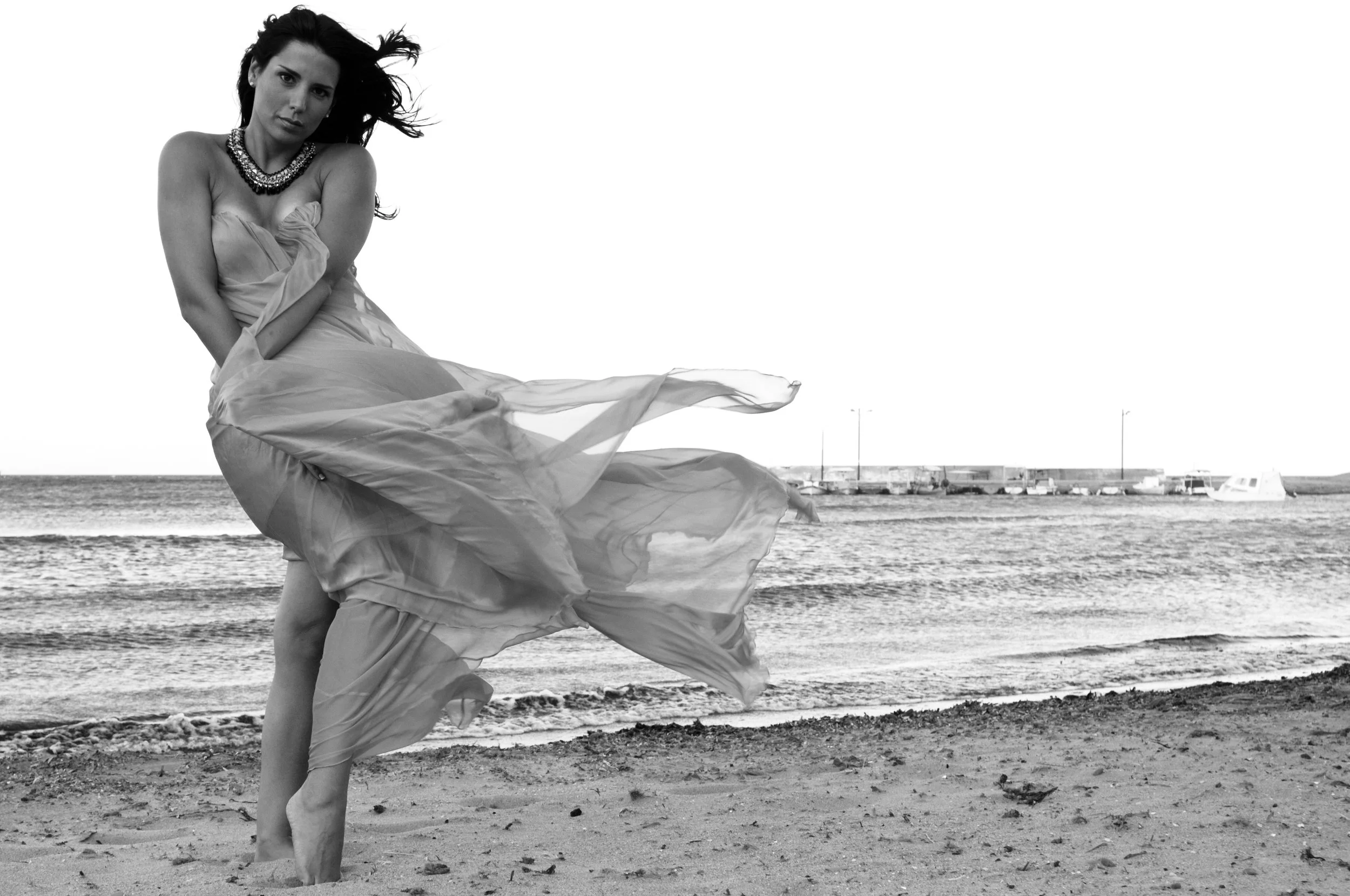 A woman in a flowing dress standing on the beach near the ocean, with wind blowing her hair and dress, in black and white