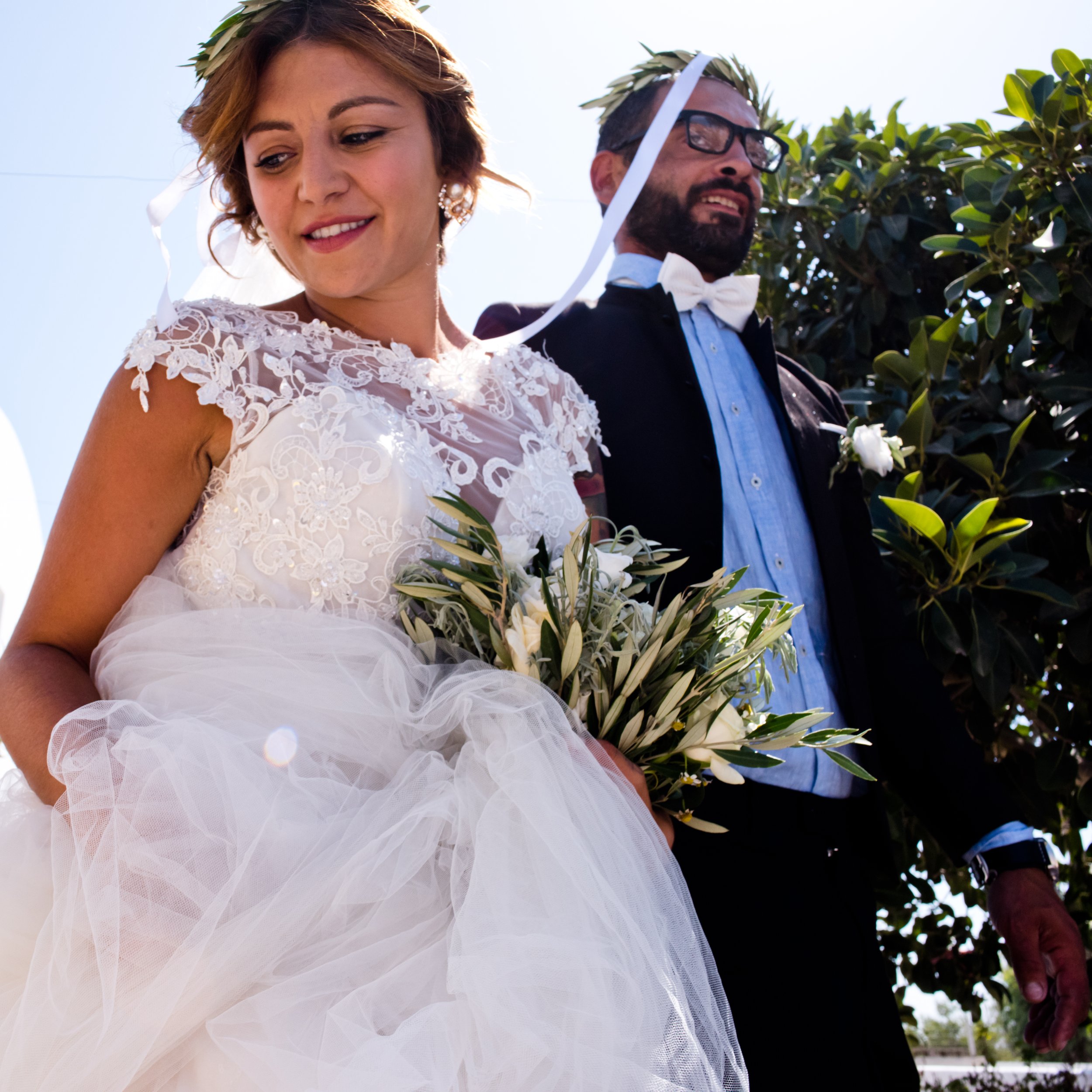 A bride and groom standing outdoors, both wearing laurel wreaths, with the bride holding a bouquet of white flowers, sunlight shining behind them, dressed in wedding attire, smiling softly.