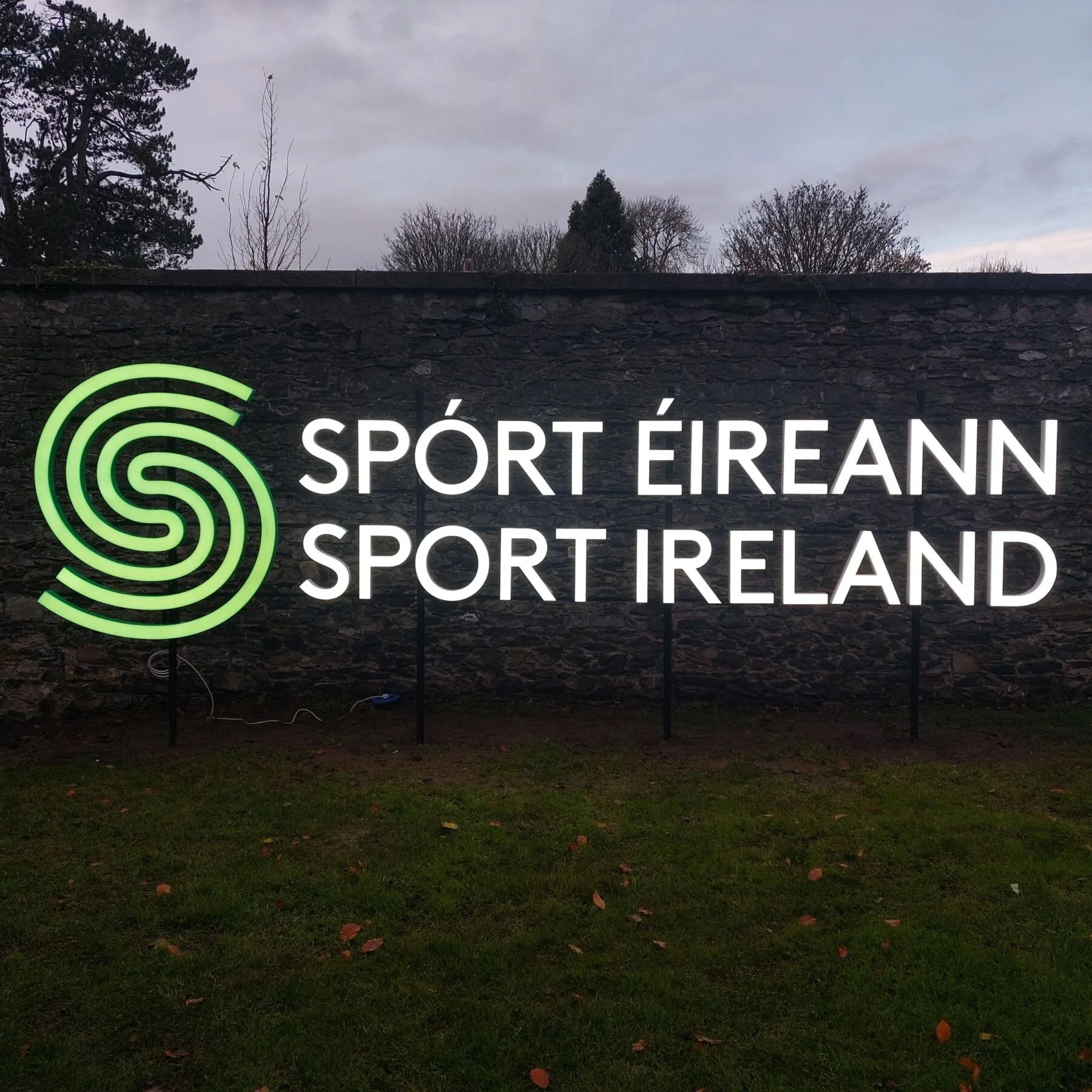 Neon sign on a brick wall with a green spiral logo and white text reading 'Sport Éireann' and 'Sport Ireland'.