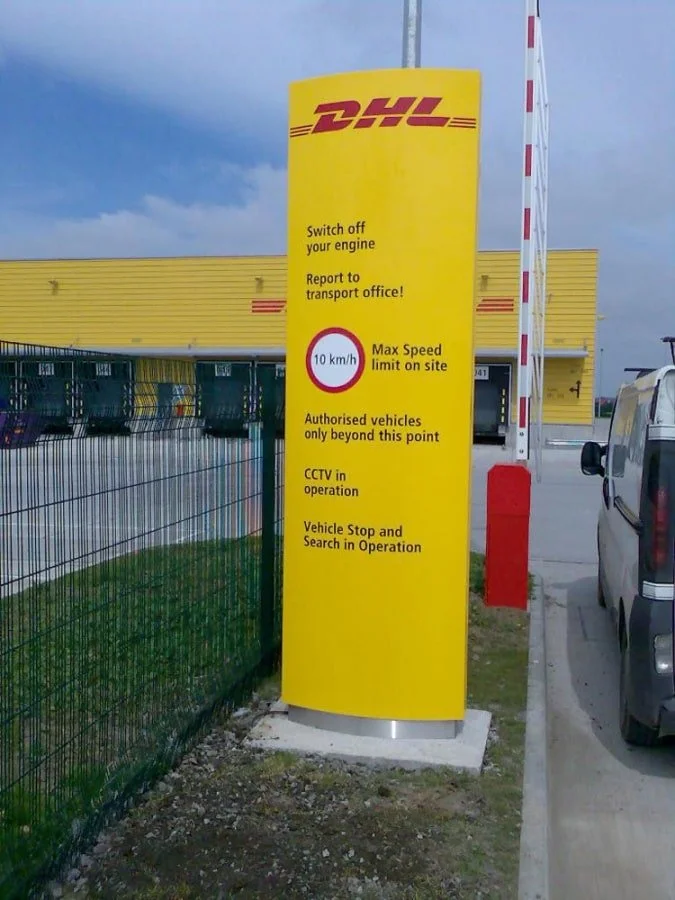 Yellow sign for DHL with safety instructions and speed limit, located next to a parked vehicle and traffic barrier.