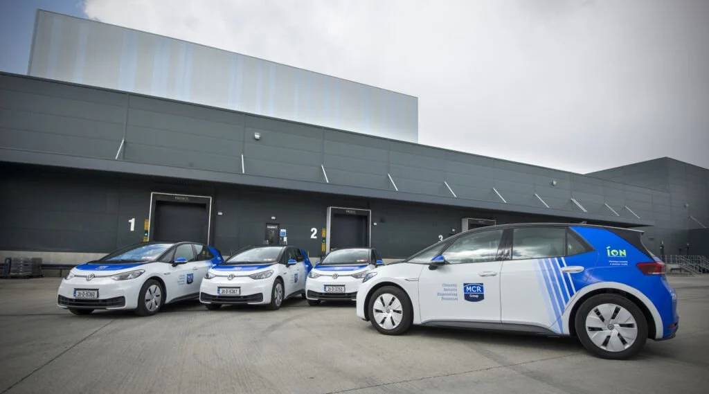 Four white and blue electric vehicles parked in front of a large industrial building with loading bays, with the third vehicle showing branding for an electric vehicle charging company.