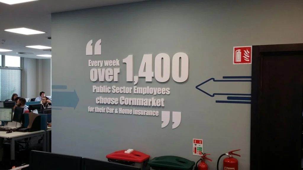 Office wall with large text stating that over 1,400 public sector employees choose Cormarket weekly for car and home insurance, with arrows and fire safety signs nearby.