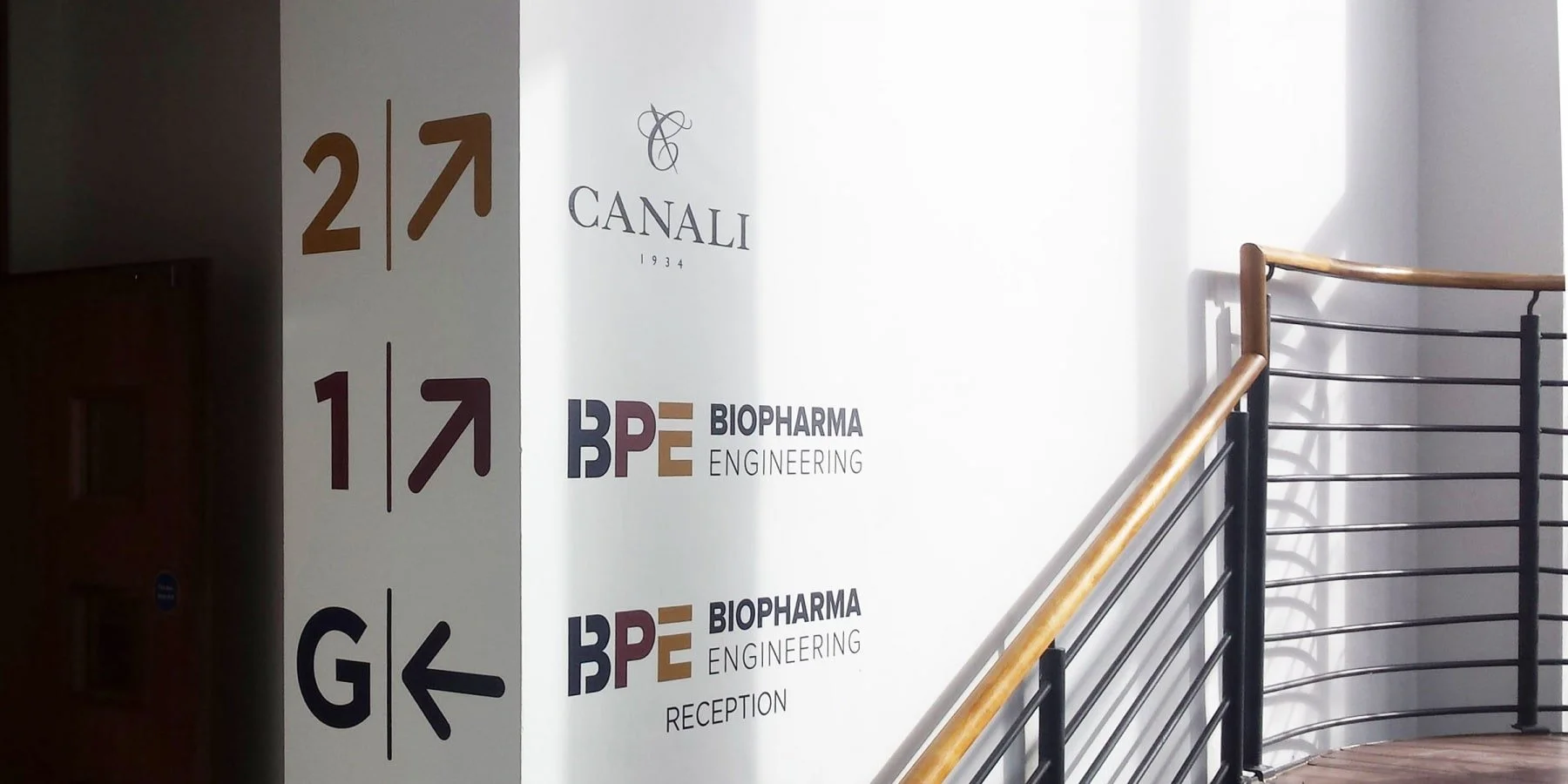 Sign with directions to rooms 1 and 2, and G and K, with logos of Canali and BPE Biopharma Engineering on the wall beside a curved railing staircase.