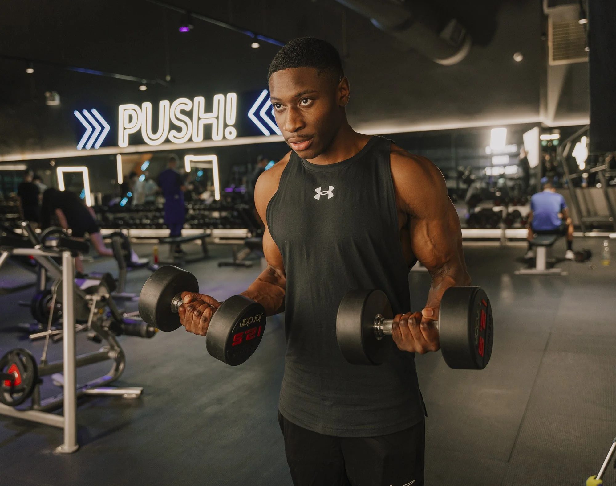 Young man lifting dumbbells in a gym with a neon sign that says 'PUSH!'