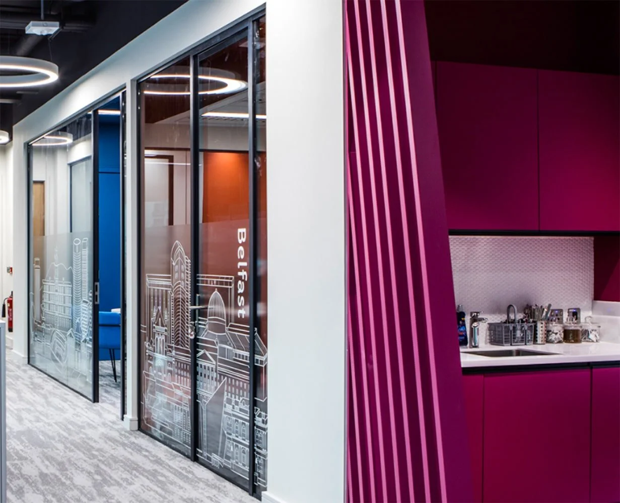 Modern office space with glass-walled meeting rooms on the left and a vibrant pink kitchenette with striped panels on the right.