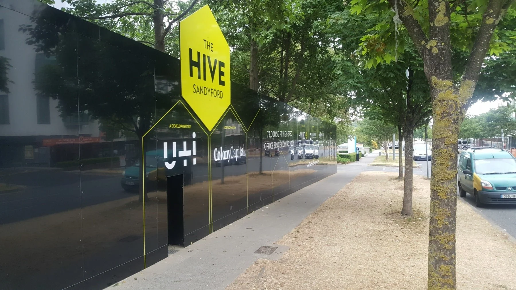 Black building wall with yellow signage for The Hive Sandovery, a development by Colony Capital, along a sidewalk with trees, parked cars, and a street in the background.