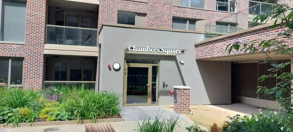 The entrance to a residential building named 'Chambers Square' with a garden in front, brick and gray concrete exterior, glass door, and an adjacent mailbox.