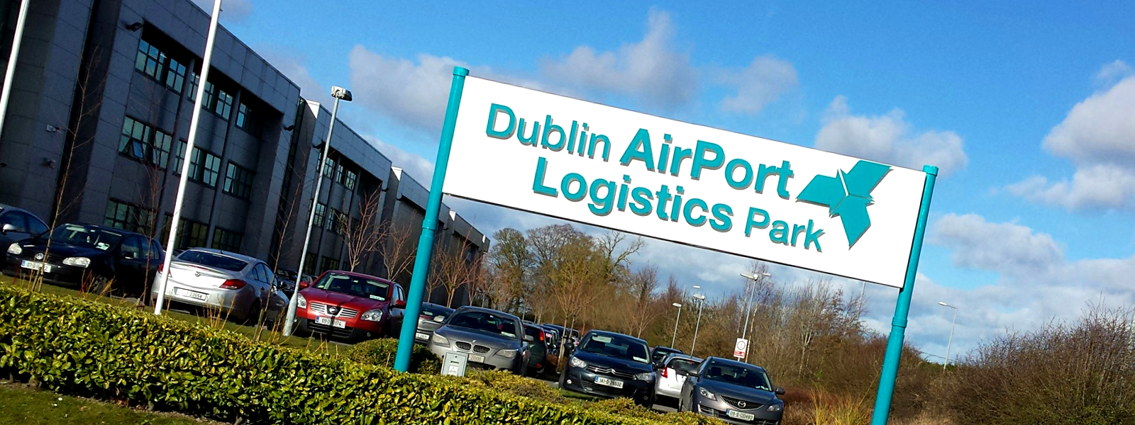 Sign for Dublin Airport Logistics Park with parked cars and a modern office building in the background under a partly cloudy sky.