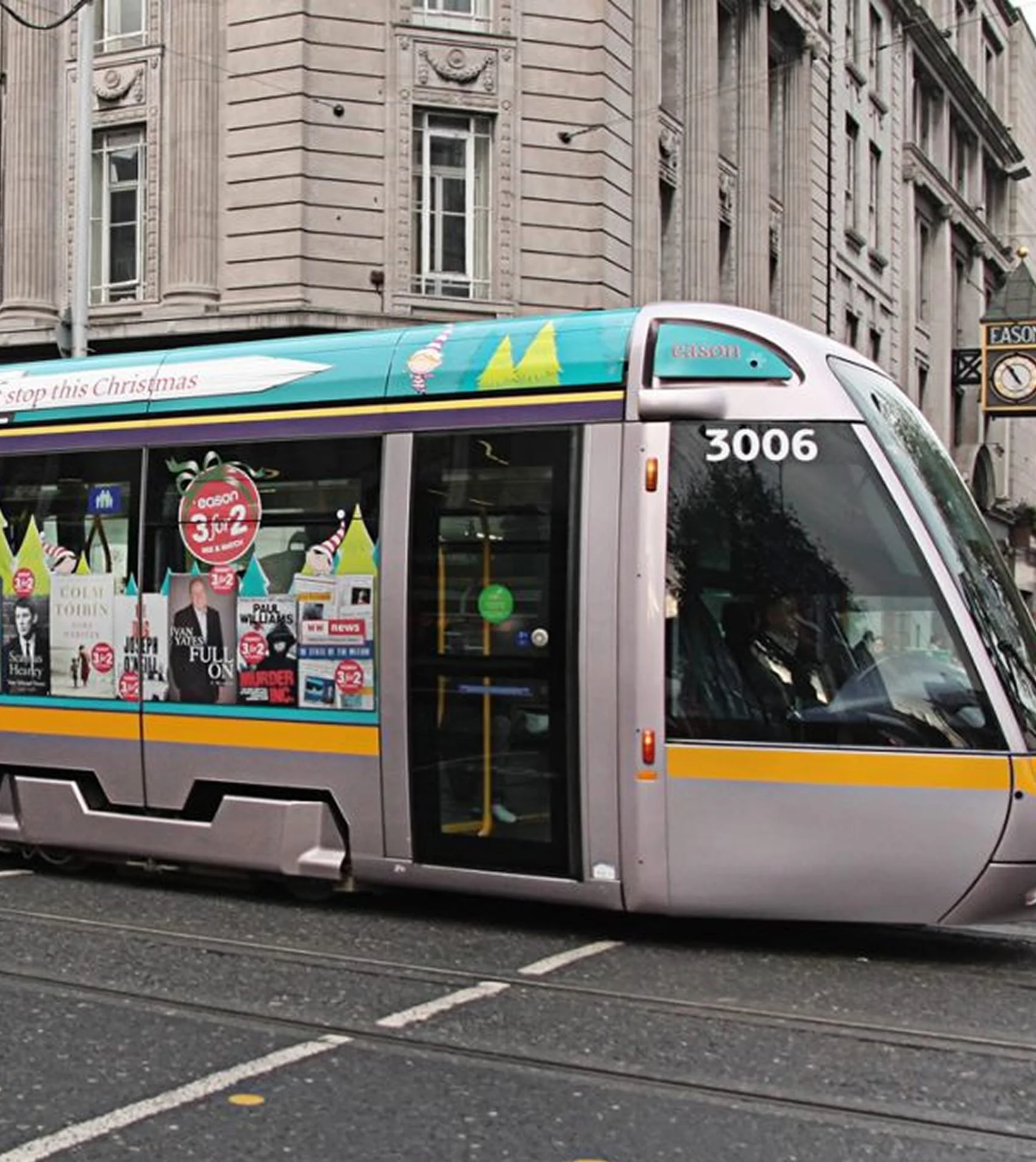 Light gray modern streetcar with advertisements and posters on the side, traveling on tram tracks in an urban area with historic buildings and a clock in the background.